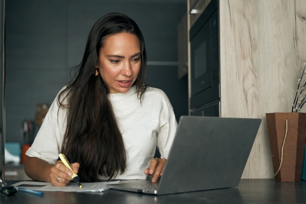 A young woman with long dark hair sits at a table annd works at a laptop. She has one hand on the laptop keyboard; the other hand uses a yellow highlighter on a piece of paper.