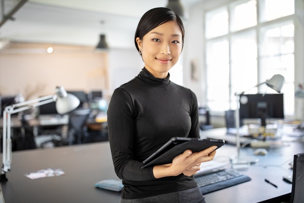  A young woman in a black turtleneck stands in a large open-plan office. She has a slight smile on her face and is holding an electronic tablet.