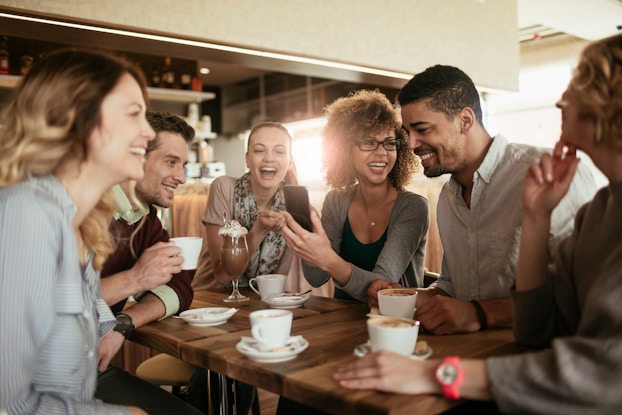 A diverse group of six friends sits around a wooden table at a warmly lit cafe. Cups of coffee and saucers are arranged on the table in front of them and the friends are all laughing as a woman in the middle of the group shows something on her smartphone to the people sitting next to her.