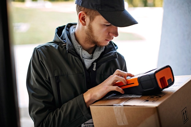  A young man wearing a windbreaker and a baseball cap examines a scanning device, which he balances on top of a package in a brown cardboard box.