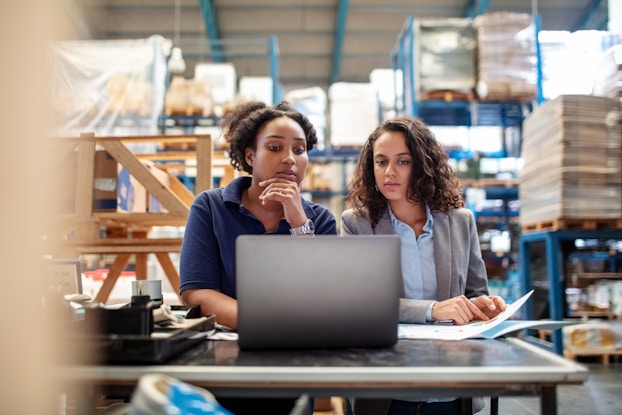  Two women stand at a table in the middle of a warehouse and look curiously at a laptop. IN the background are shelves filled with pallets of goods wrapped in plastic.