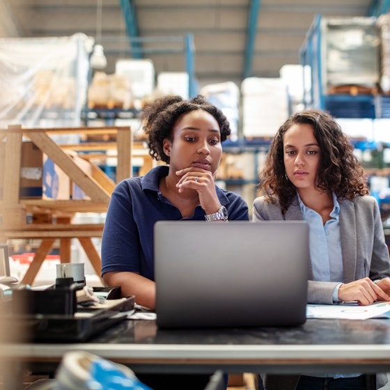  Two women stand at a table in the middle of a warehouse and look curiously at a laptop. IN the background are shelves filled with pallets of goods wrapped in plastic. 