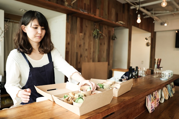  A woman wearing a dark blue apron stands behind a long wooden counter and packs food into a recyclable cardboard takeout box. The box is filled with small cardboard cups of sliced green vegetables and rice. An identical box, already filled, sits next to the first box on the counter.