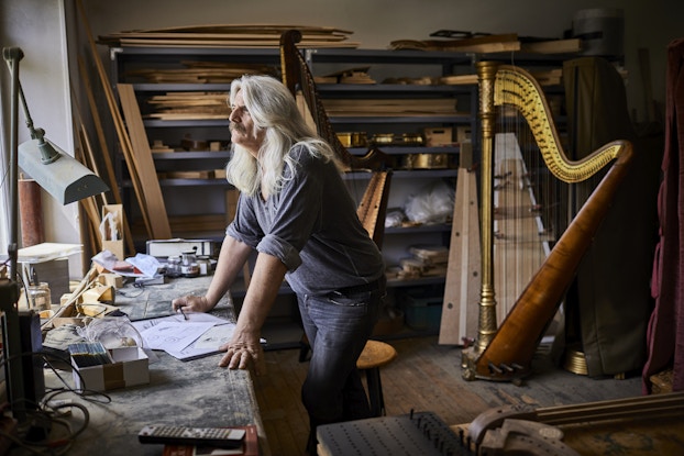  An older man stands in profile in a workshop, leaning on a cluttered desk and staring out an unseen window. The man has long silver hair and a gray mustache, and he wears a dark gray shirt with the sleeves rolled up to expose his forearms. In the background, one wall of the workshop is filled with shelves of pieces of wood. A handmade musical harp, almost as tall as the man, leans against the shelves.