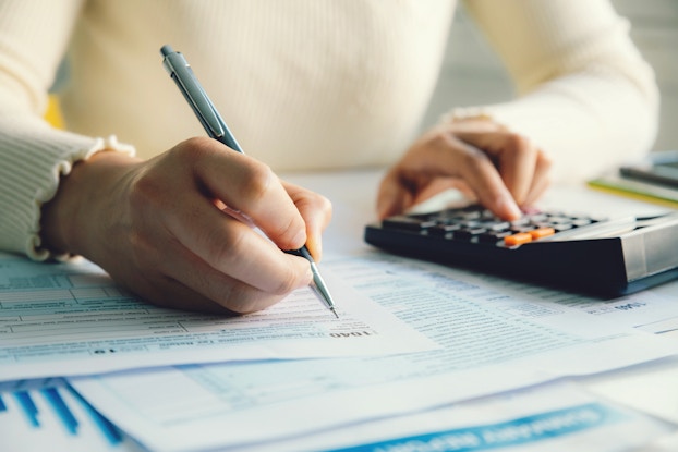 Close-up on a pair of hands. One hand uses a mechanical pencil to fill out a form while the other presses buttons on a calculator.