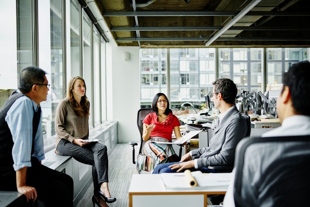  A group of five office professionals, including men and women, are seated in a circle and discuss a work project. The background is a modern office building.