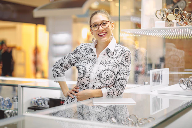 A woman owner of a small jewelry stores smiles at the viewer. She is leaning against a case that is displaying fine watches and jewelry.