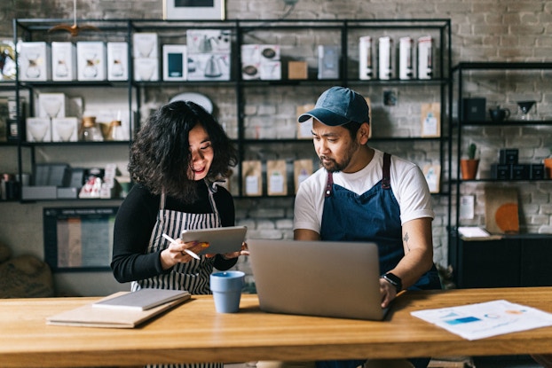 Two co-owners of a coffee shop are seated at a table and are engaged in a lively discussion. They are reviewing data on a tablet and a laptop.