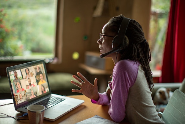 A woman wearing an over-the-ear headset sits at a table in front of an open laptop and speaks into the headset's mouthpiece. The laptop screen is split into nine boxes, each of which shows a different person. In the background, out of focus, can be seen the woman's home, with a window seat beneath a window and a dog curled up on a chair.