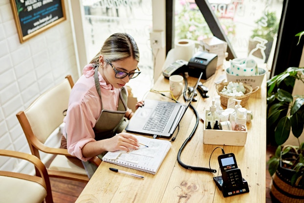  A woman wearing eyeglasses and an apron over casual clothing sits at desk in her shop doing bookkeeping.