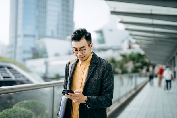 A man standing on an elevated walkway looks down at his smartphone. In the background are several monochrome city buildings, including a mirrored skyscraper, and clusters of trees.