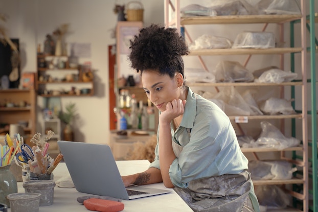 A woman sits at a counter in front of an open laptop with one hand on her chin in thought. The room around her is an artist's workshop, with shelves of sculptures wrapped in plastic in the background and cups of carving and drawing tools clustered on the counter. The woman wears a loose light blue shirt and she has a gray clay-smeared apron wrapped around her waist.