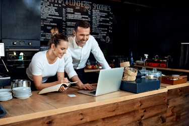  A man and a woman stand behind the counter of a cafe and lean forward to look at the screen of the laptop sitting open on the countertop. The woman is writing something in a notebook, and both the man and the woman are smiling. 