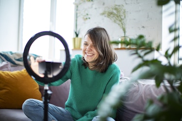 Person sitting on couch in living room smiling at a phone placed in the center of a ring light, recording a video. 