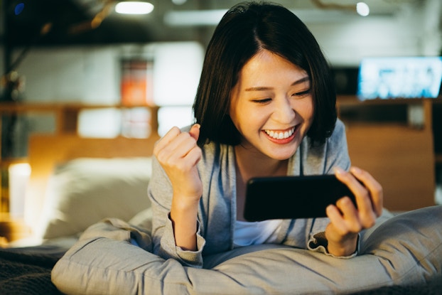  A young woman lays on her stomach on a light gray pillow and looks at the smartphone held horizontally in her hand. She smiles and pumps her fist at what she sees on the screen.
