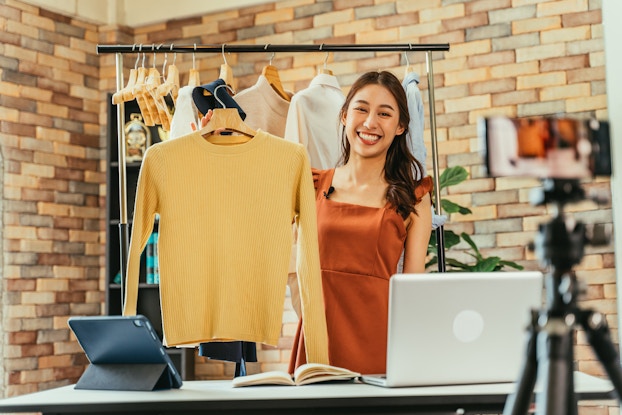  A woman in an orange dress holds up a yellow long-sleeved shirt as a smartphone on a tripod films her. Standing between the woman and the smartphone is a desk holding an open book, an open laptop, and a smart tablet on a stand. In the background is a rack hung with several other shirts.