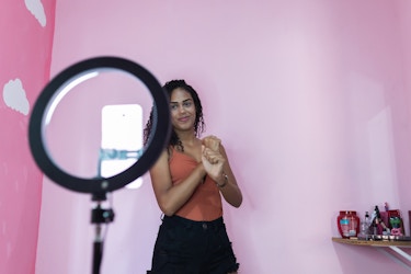  Woman posing for a phone with a ring light in front of a pink background. 