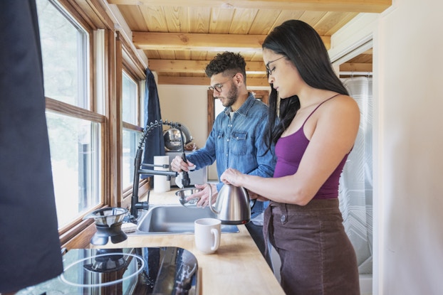  A couple seen in profile stand in front of the sink in the kitchen area of their tiny home. The person closest to the viewer is a woman with long dark hair; she wears glasses and a purple tank top, and she holds a small silver teapot above a mug, getting ready to pour a cup of tea. The man next to her has curly hair and a beard; he wears glasses and a blue button-up shirt. He uses the flexible and detatchible sink faucet to wash out a silver bowl. The kitchen area is a countertop holding a few appliances. Two windows over the countertop look outside. Behind the couple is an open doorway, through which can be seen a shower.