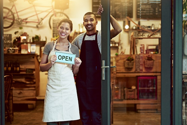 A man and a woman stand smiling in the doorway of a shop. The woman holds an OPEN sign.
