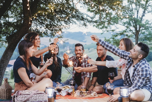  A group of seven friends sit on an orange picnic blanket outside, with a scenic valley in the background and trees strung with lights overhead. In front of the group are glasses of wine, a couple of lanterns, a charcuterie board, and mason jars filled with a golden beverage. Each member of the group holds up a lit sparkler.