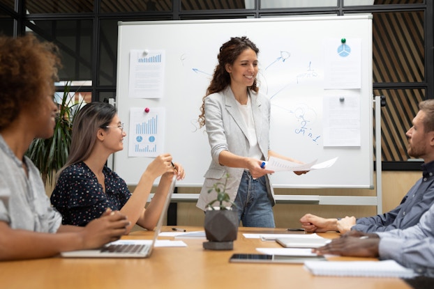  woman presenting to coworkers in a meeting in front of a whiteboard