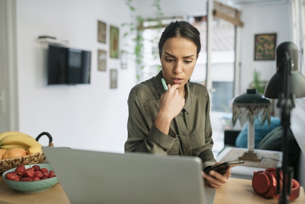 A woman sitting in her home looks thoughtfully at her laptop. In one hand is a pen and in the other is a smartphone.