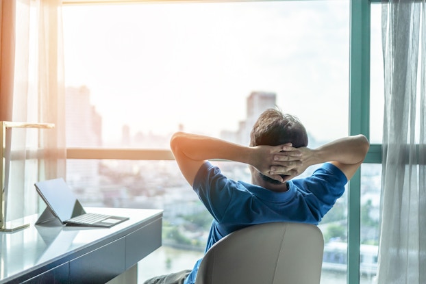 A man leans back in a chair with his hands behind his head and looks through a floor-to-ceiling window at a sprawling cityscape. At the man's left is a sleek modern desk with a laptop sitting abandoned.