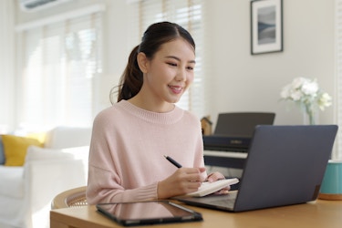  A young woman with her hair in a ponytail sits at a table in front of an open laptop. She takes notes on a notepad while watching something on the laptop's screen. In the background, out of focus, are a white couch, a piano keyboard and a tall plant with white flowers. 