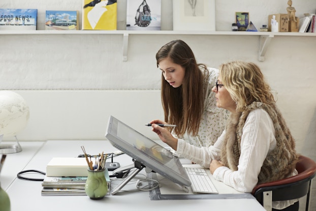 A woman, seen in profile, sits at a white table in front of a digital drawing tablet and a computer keyboard. She wears a fur vest over a white sweater and holds a digital stylus or pen in one hand, as if getting ready to draw something on the tablet. Another woman stands over her, leaning forward to look at the tablet. On the wall behind the two women, a high shelf holds small canvases of artwork.