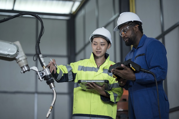  Two people in white hard hats stand in front of a robotic arm that ends in a welding tool. The person on the left, a woman in a neon yellow high-visibility shirt, is using one hand to indicate something on the welding arm and the other hand to hold an electronic tablet. The person on the right is a man in a blue jumpsuit and safety goggles. He holds a black control panel connected to a cord.