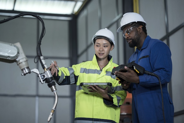 Two people in white hard hats stand in front of a robotic arm that ends in a welding tool. The person on the left, a woman in a neon yellow high-visibility shirt, is using one hand to indicate something on the welding arm and the other hand to hold an electronic tablet. The person on the right is a man in a blue jumpsuit and safety goggles. He holds a black control panel connected to a cord.