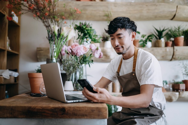  A young man in a brown apron sits at a wood-topped counter and looks at the smartphone in his hand. Next to him on the counter are an open laptop and several bouquets in vases. In the background are several shelves designed to look as if they're made of halved tree trunks. The shelves hold various potted plants.