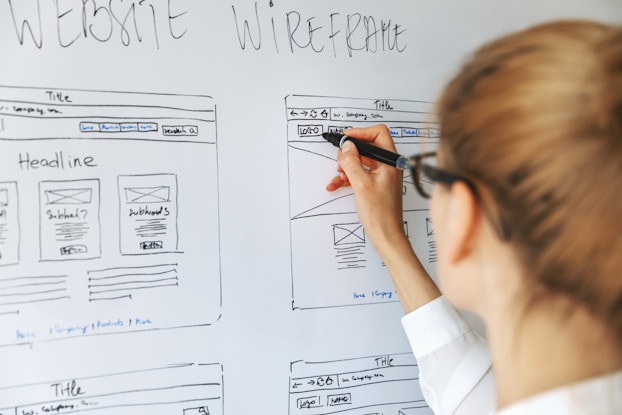  A woman seen from behind adds to a drawing on a whiteboard. The drawings show various pages of a website and their layouts under the heading "WEBSITE WIREFRAME."