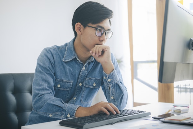  A young man in glasses and a denim shirt sits in a padded office chair and works on a computer. One of his hands is on the keyboard and the other hand is pressed to his chin as he considers the monitor in front of him.