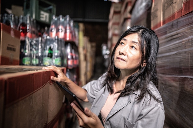A mature woman works in a storage room checking the inventory and entering it on a tablet computer.