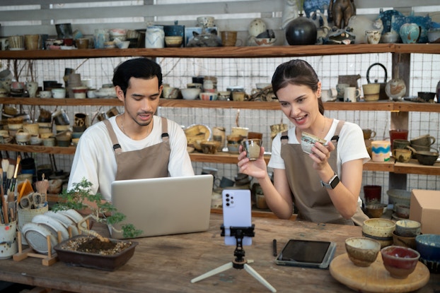 Two ceramics artists sit side-by-side at a wooden counter facing a smartphone on a tripod, which sits on the countertop. Both artists wear white T-shirt and brown aprons. The artist on the right is a dark-haired woman who holds two ceramic teacups and speaks to the filming phone. The artist on the left is a dark-haired man with a mustache and beard; he works on a laptop. Behind the two artists are shelves filled with ceramic cups, bowls, vases, and assorted other works of art.
