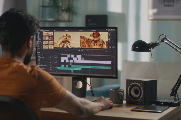  An over-the-shoulder shot of a man in an orange shirt working on video-editing software on a computer monitor. The monitor shows two still shots showing different angles of an astronaut in a reddish rocky landscape. Below the still shots are several purple and green bars showing where video has been cut and inserted.