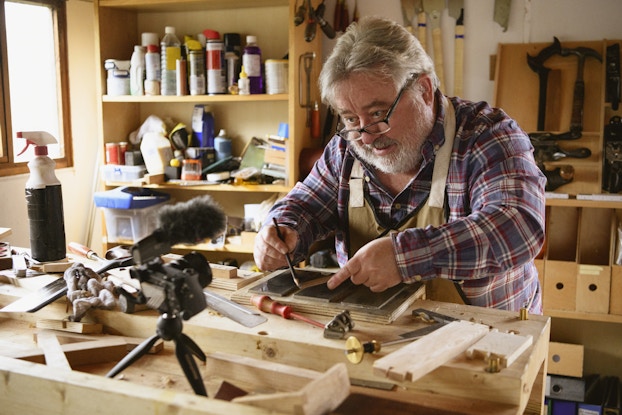 A bearded gray-haired man works on a wood-working project in front of a small camera and boom mic mounted on a tripod. The man uses a pencil to trace the shape of a smaller plank of wood onto a larger plank of wood. He wears a red-and-blue plaid shirt under a khaki apron and is looking at the camera over the tops of the glasses perched near the end of his nose. The man's workroom is cluttered; there are shelves behind him crowded with various spray bottles, and the worktable is scattered with various tools and pieces of unstained wood.