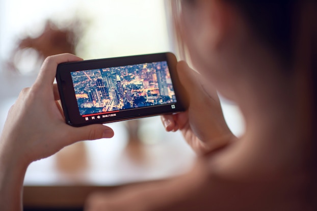  An over-the-shoulder shot of a person watching a video on a smartphone. The person holds the phone horizontally in both hands. The red bar at the bottom of the phone's screen shows that the video is about 1/3 completed, and the video shows a city from above with its buildings illuminated in the night.