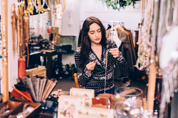 A woman stands in the workroom of a jewelry shop, stringing a gold necklace. The woman has long brown hair and wears a black Fair Isle-style sweater with white snowflake patterning. The room and its worktable are cluttered with necklaces and other jewelry pieces hanging from stands and hooks, a rack of coats, shallow wooden boxes filled with jewelry, and folders of dcouments.