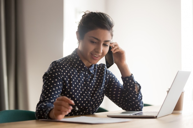 A woman sits at a table in front of an open laptop with a smartphone pressed to one ear. She speaks into the phone as she writes something on a piece of paper.