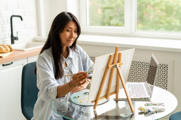 A woman sits at a small round table and uses a thin paintbrush to apply paint to a square canvas. The canvas is held on a small wooden easel that sits on top of the table, next to an open laptop, a palette spread with a few different colors of paint, and three tubes of paint.