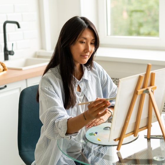 A woman sits at a small round table and uses a thin paintbrush to apply paint to a square canvas. The canvas is held on a small wooden easel that sits on top of the table, next to an open laptop, a palette spread with a few different colors of paint, and three tubes of paint.