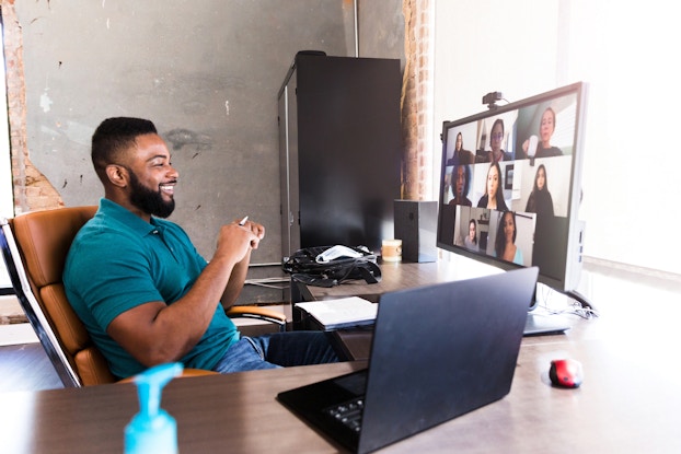 A man sits at a desk facing a large monitor with a webcam. On the monitor are video feeds of several other people in a meeting.