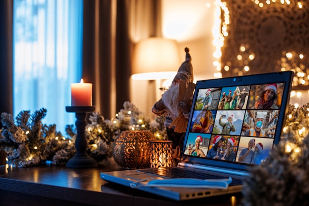 A table in a warmly lit room. A laptop screen shows a three-by-three grid of nine video feeds showing various people in various locations. Most of the people on-screen are wearing Santa hats; some are outside in tropical locations or in front of a house. Around the laptop, the table is decorated with pine garland, lit candles and a small statue of a bearded elf in a Nordic-style hat and sweater.