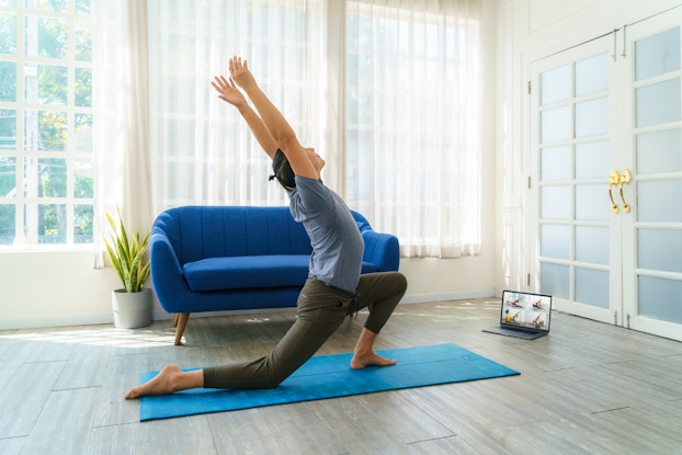 A man crouches on a turquoise yoga mat with one knee on the ground and his arms stretched upward. On the screen of the open laptop in front of him, four other people, each in their own window of a video chat program, make similar poses.