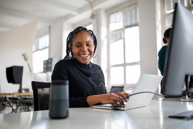  A smiling businesswoman seated at a table multitasks. She is smiling as she uses a smart speaker while working at her desk in office.