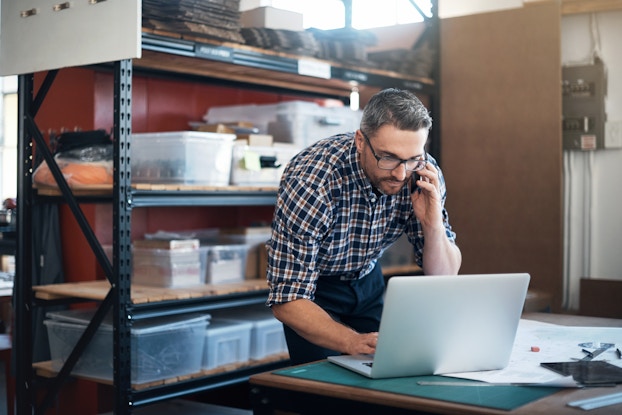 A man with salt-and-pepper hair leans over an open laptop. He uses one hand to type something and the other hand to hold a smartphone to his ear. The man wears glasses and a black-and-white plaid shirt. Behind him is a metal shelving unit holding many large plastic bins and boxes.