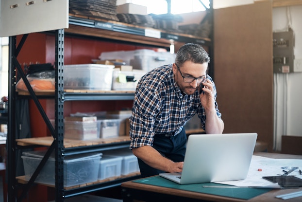  A man with salt-and-pepper hair leans over an open laptop. He uses one hand to type something and the other hand to hold a smartphone to his ear. The man wears glasses and a black-and-white plaid shirt. Behind him is a metal shelving unit holding many large plastic bins and boxes.