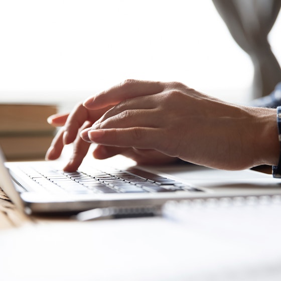 A close-up of someone's hands typing on a laptop keyboard. Most of the person in the picture is out-of-frame, but it can be seen that they are wearing a dark blue shirt with white polka dots.